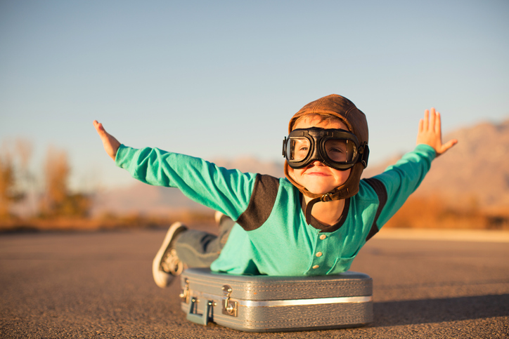 A young boy with outstretched arms lies on top of a suitcase imagining he is flying on an airplane away and traveling to exotic locations. He is wearing a flight cap and goggles and has a large smile on his face. He loves to travel.